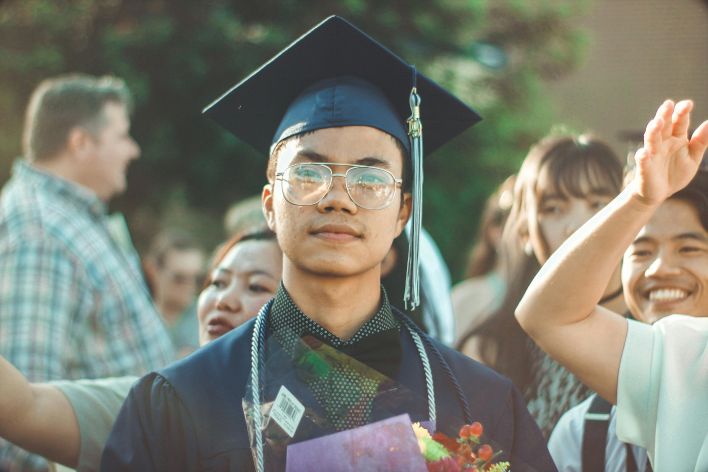 Asian male graduate smiling during an outdoor graduation celebration with family and friends.
