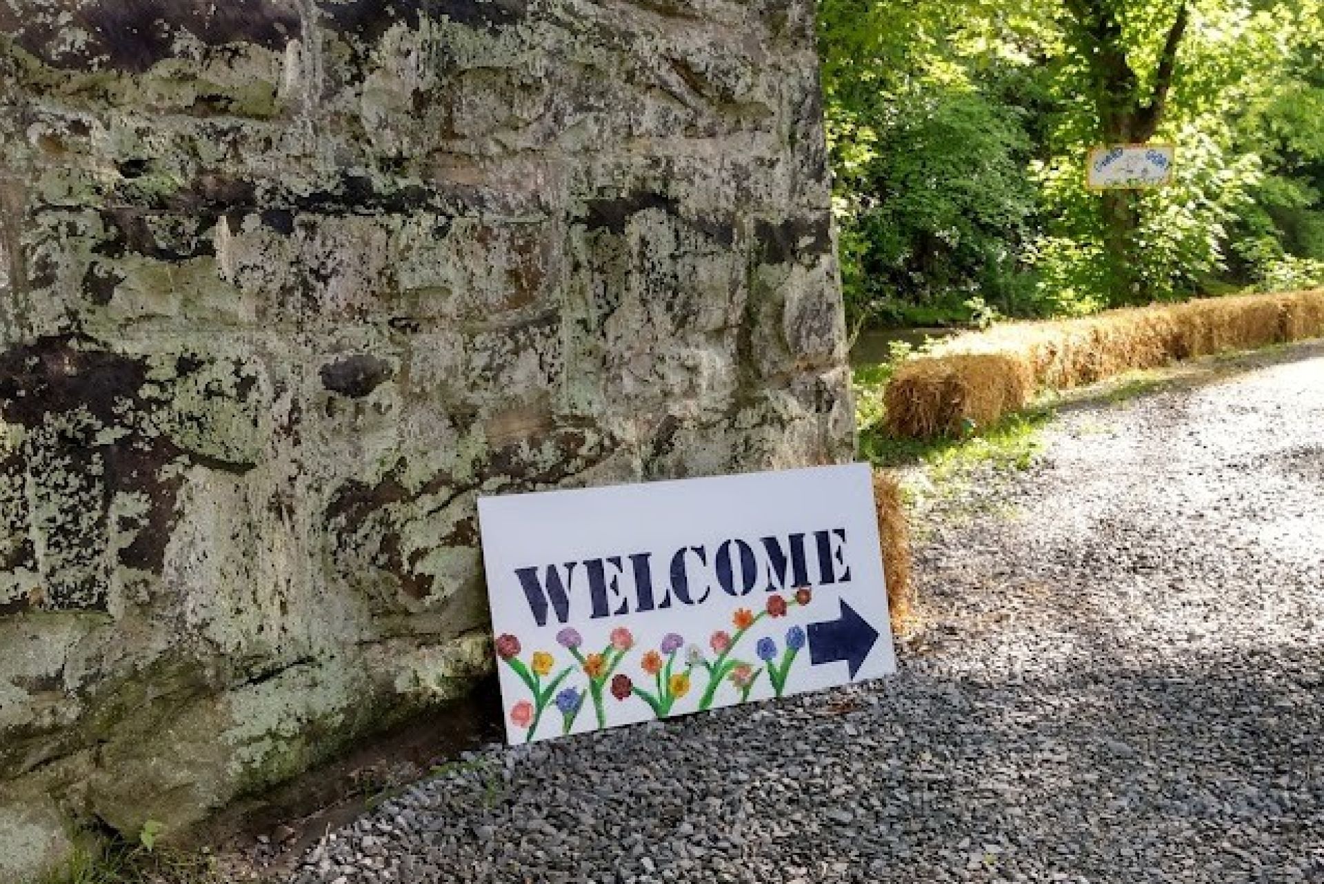A welcome sign resting against a stone wall with a sunlit trail lined by bales of hay.